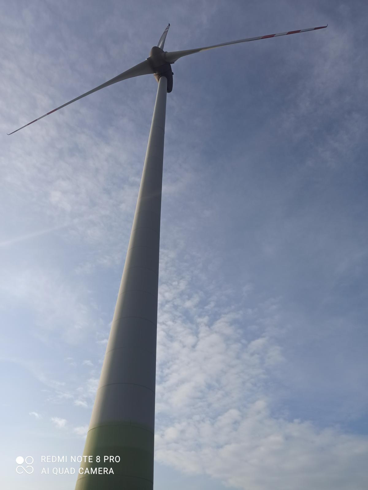 Niski kąt widzenia na turbinę wiatrową z trzema łopatami na tle błękitnego nieba z chmurami.